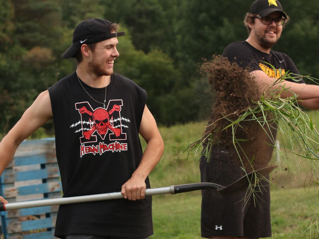 Two students from the University of Waterloo volunteering on Our Farm. Student on left is tossing soil in the air with a shovel while smiling. Student behind on the right is watching them and smiling.