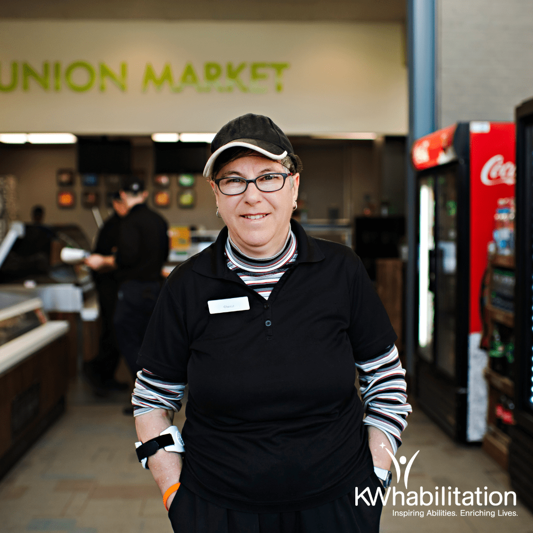 Person standing outside of Union Market in a work uniform smiling. (KW Habilitation logo on bottom right)