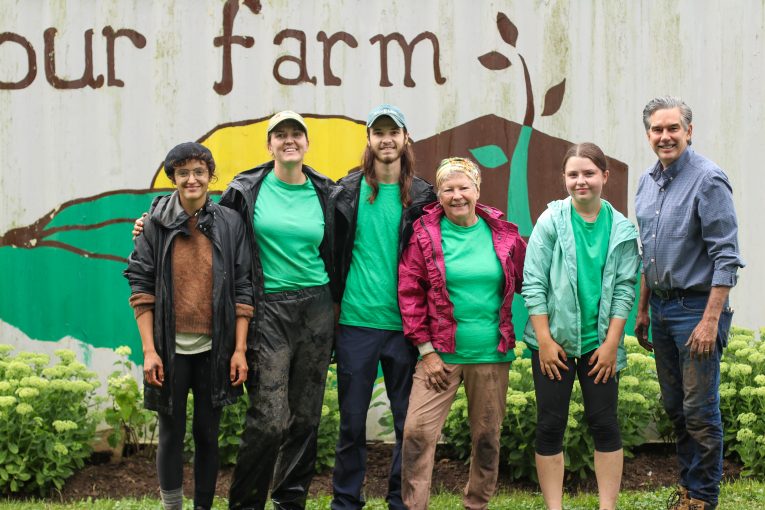 (Standing from left to right) Dani a CSJ staff, Laura Bredschneider an Our Farm farmer, Aidan a CSJ staff, Our Farm Coordinator Jenny Weickert, Evie a CSJ staff, and MP Tim Louis all smiling in front of the Our Farm mural.