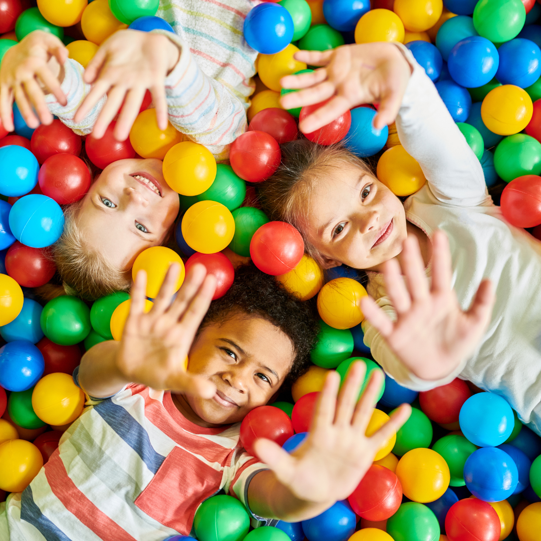 three kids lying in a ball pit, the photo is taken from above and there are bright coloured balls all around them. Their hands are in the air and they are smiling.