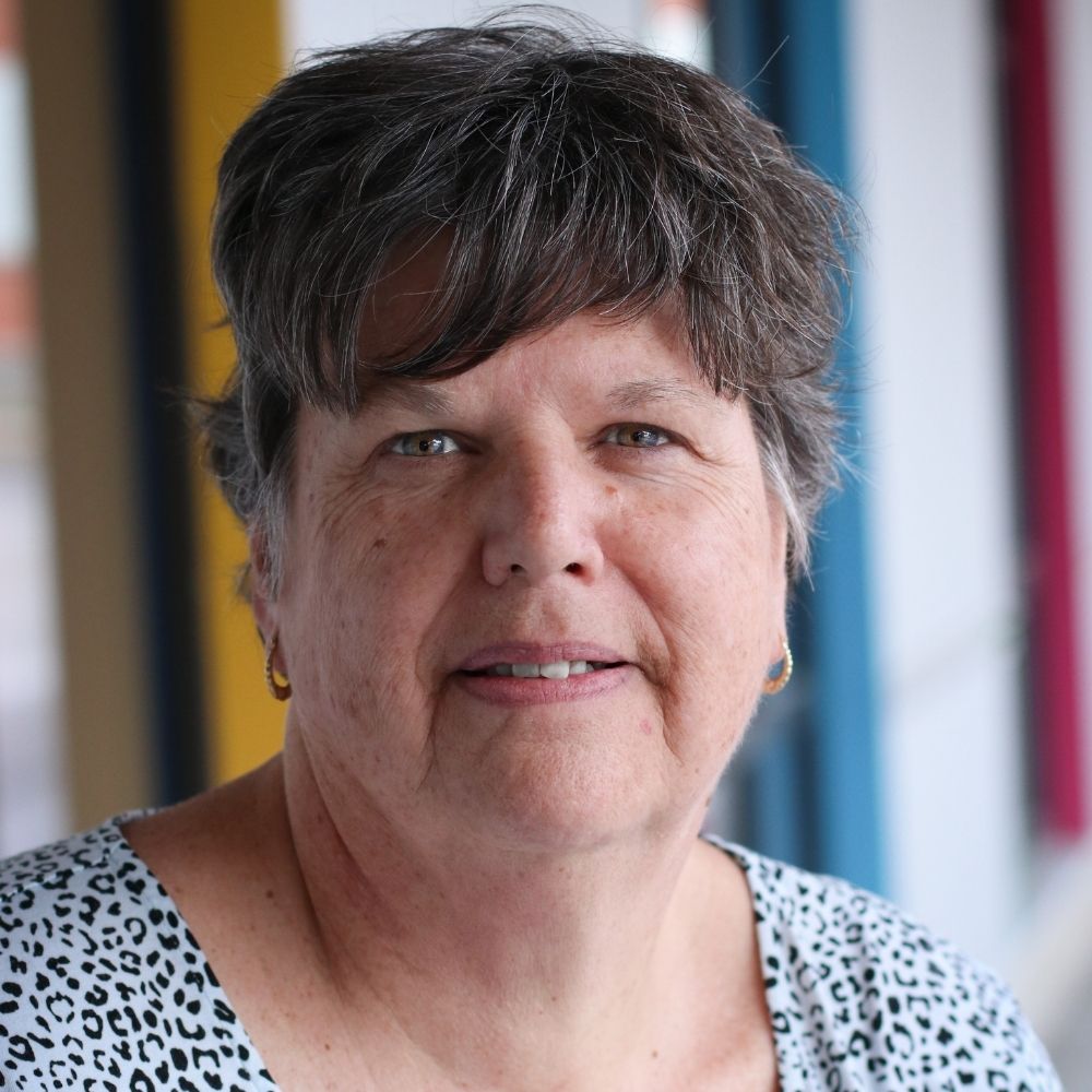 headshot of a woman with brown and grey hair, blue eyes wearing a polka dot shirt.