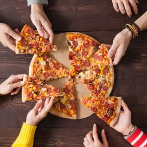 a group of people sharing a pizza. the photo is from overhead and focuses on a pizza with lots of hands coming in from the sides grabbing a slice.