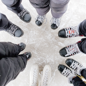 6 people standing in a circle looking down at their ice skates on the ice.