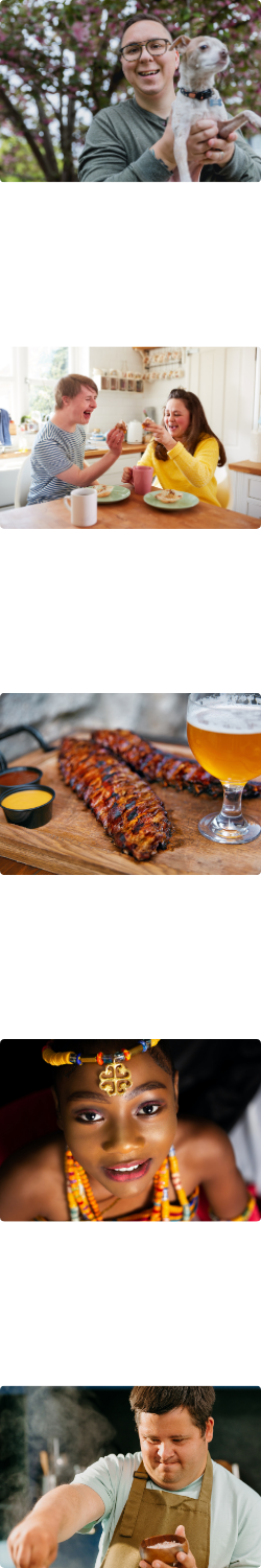 A person holding a dog. Two people enjoying eating. A rack of ribs and a glass of beer. A person wearing African beading. A person adding seasoning when cooking.
