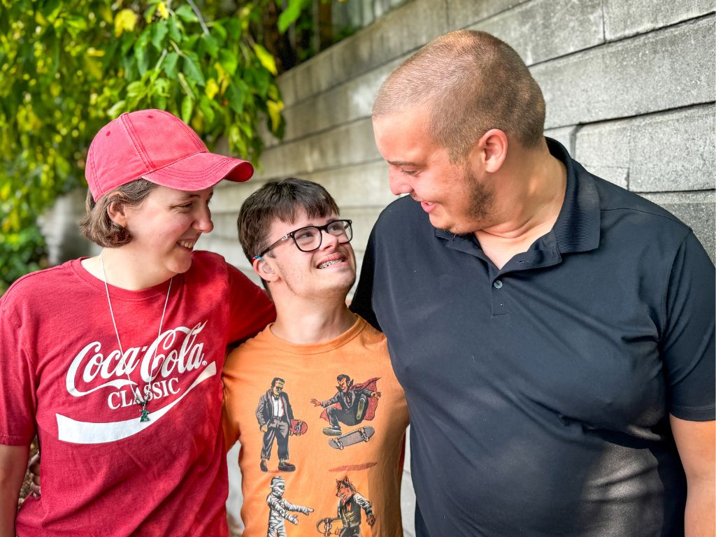 Three young people with disabilities standing by a wall with some greenery in the background,. They have their arms around each other and they're smiling and looking at each other.