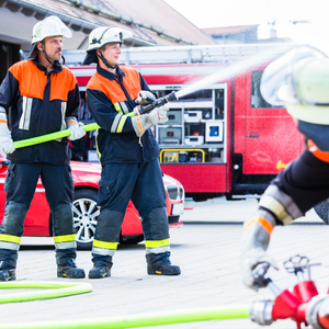 Two fire fighters holding a hose that is spraying water while another firefighter is turning a valve on a hose line.