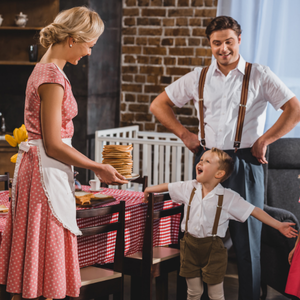 A family with a mom, dad and son dressed in old fashioned clothes. The mom is holding a stack of pancakes and the son is smiling and excited.