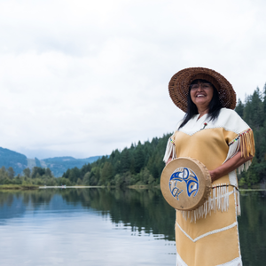 A woman wearing indigenous style clothing and holding a drum. She is smiling and standing next to a calm lake with trees around it.