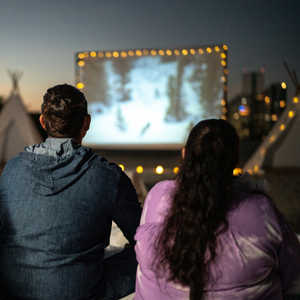 Two people dressed in warm clothing watching a movie on a big white screen outdoors.