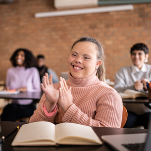 A person in a classroom with a book in front of them. They are smiling and clapping.