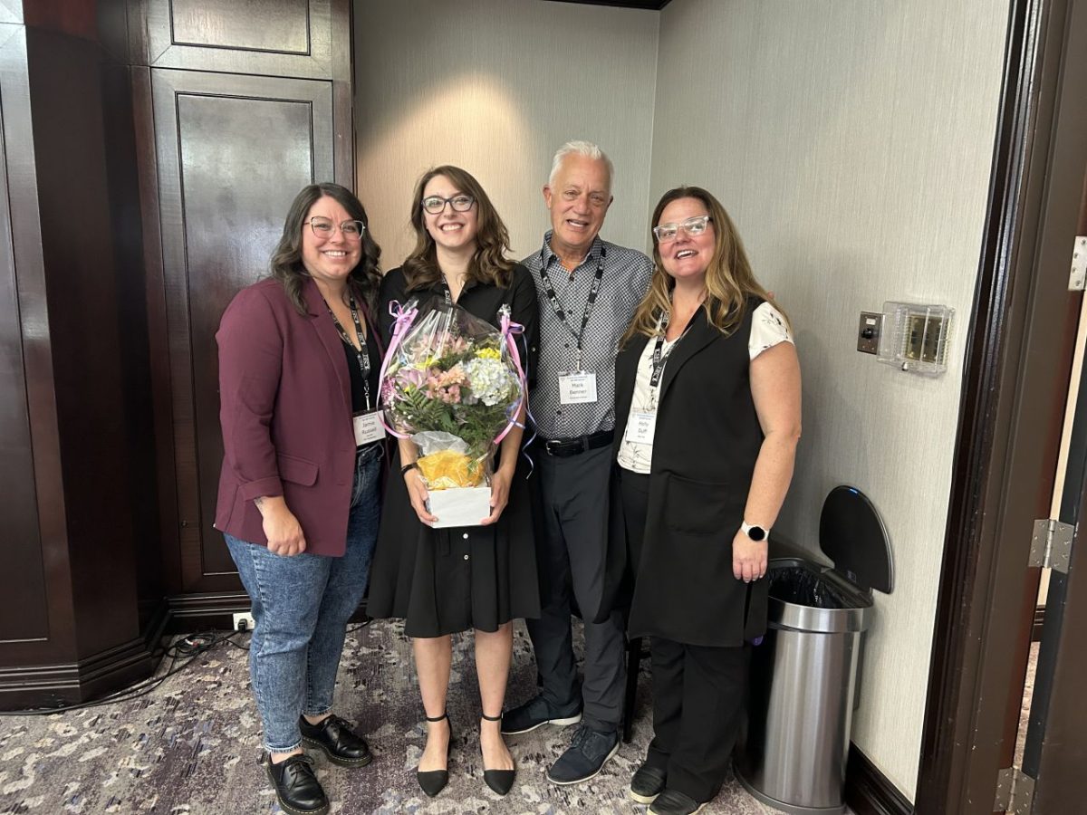 group photo of four people. One woman is holding flowers and has been congratulated by the other people in the photo. They're all smiling and happy.