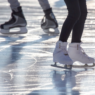 the legs and skates of two people on an outdoor skating rink. The sun is casting long shadows as it is likely in the evening.