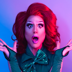 A drag queen with bold eye makeup, a wig and a sparkly outfit with a surprised face.
