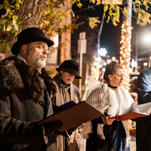 A group of people wearing warm coats and scarves with books of sheet music, singing outdoors.