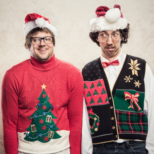 Two people looking goofy with some festival holiday sweaters. The person on the left has a christmas tree on theirs. The person on the right has a red bowtie on with a white shirt and a christmasy sweater vest with different patterns on it including trees, stars, a present and plaid.