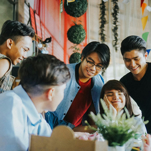 a group of five young people hanging out. There are some plants hanging and on table they are gathered around.