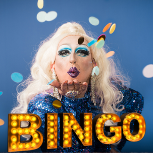 A drag queen with bold makeup wearing a blue sparkling dress. They are blowing round circles scattered through the air. There is a sign made of yellow lights that says "Bingo"