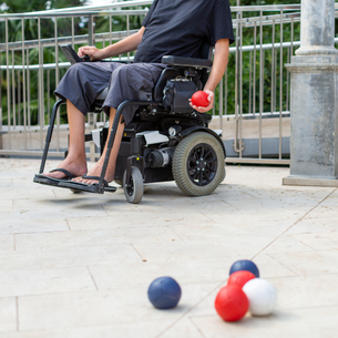 A person using a wheelchair with a red ball in their hand. It looks like they are going to bowl the ball on the ground There are several blue and balls bunched together on the ground in front of the person near a single white ball.