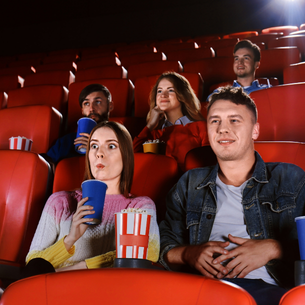 People sitting in a movie theatre with red seats watching a movie, eating popcorn and drinking beverages.