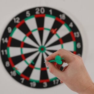 A person's hand holding a green dart. There is a dart board in the background.
