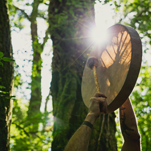 A person holding a drum up in the air and hitting it with a stick that has a ball on the end of it. There are trees and sunshine in the background.