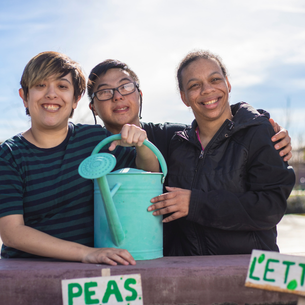 Three people standing outdoors, smiling. One has a watering can. There are two signs on the table in front of the three people. One says "peas" and the other sign says "lettuce"
