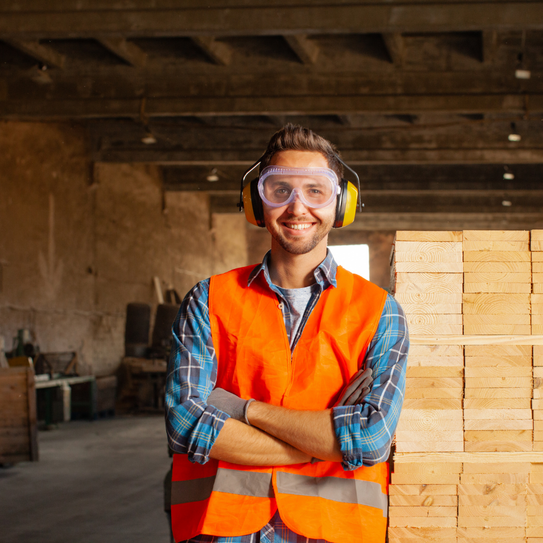 man in a high visibility vest, safety goggles and ear protection smiling and standing in front of a bunch of wood in a tidy pile