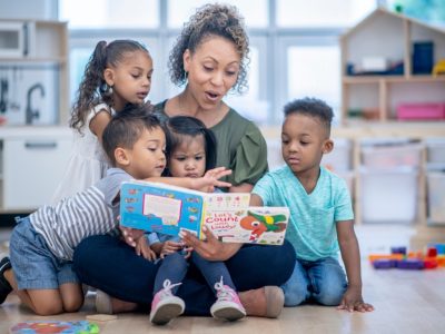 A teacher sitting on the floor of a classroom reading a book to 4 kids.